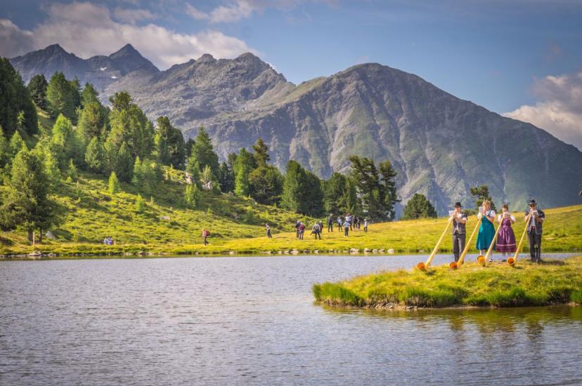 Landscape with lake, mountains and trees with three people playing traditional alpine horns