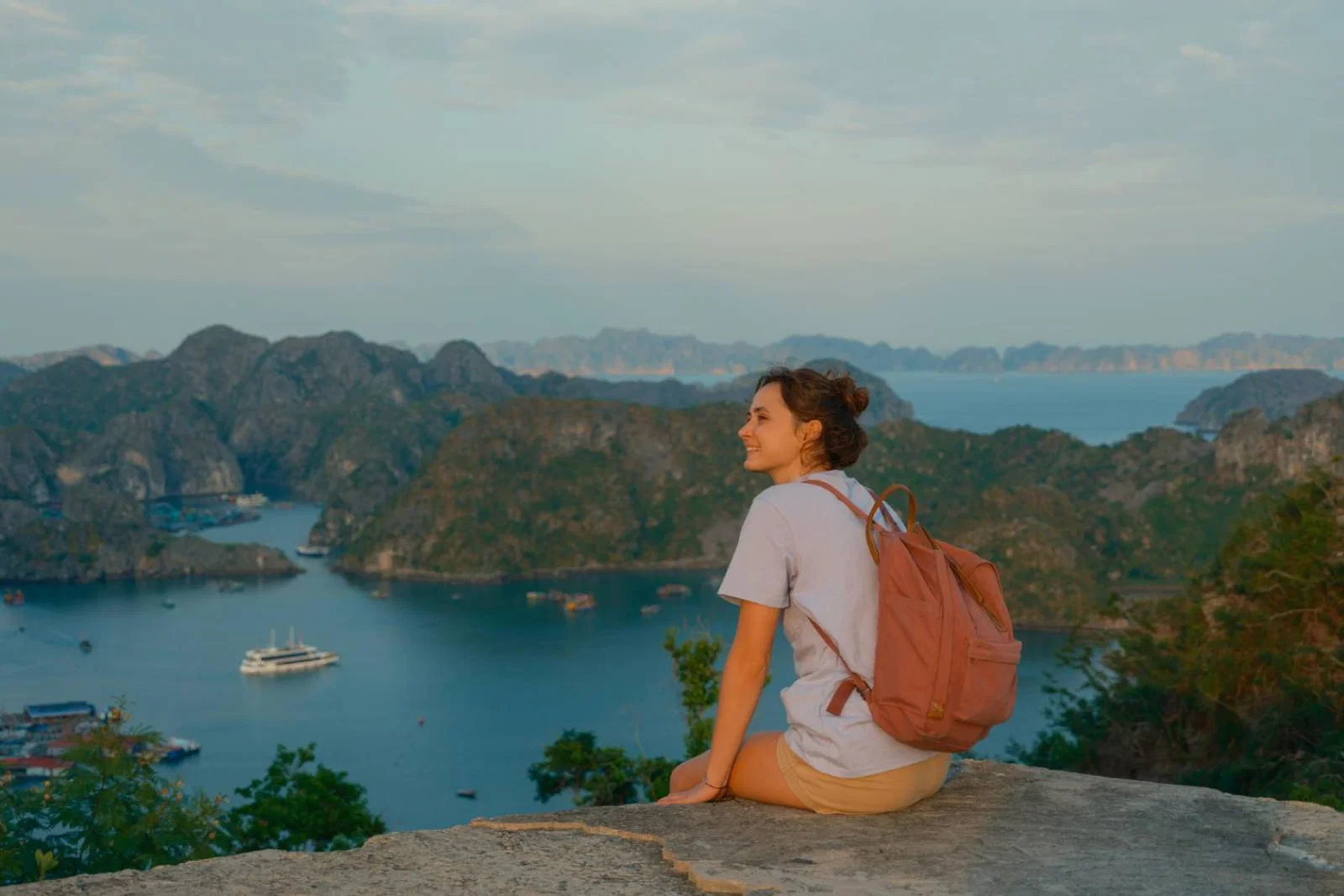 Brunette female solo traveller with backpack sitting on cliff edge overlooking scenic Vietnamese bay