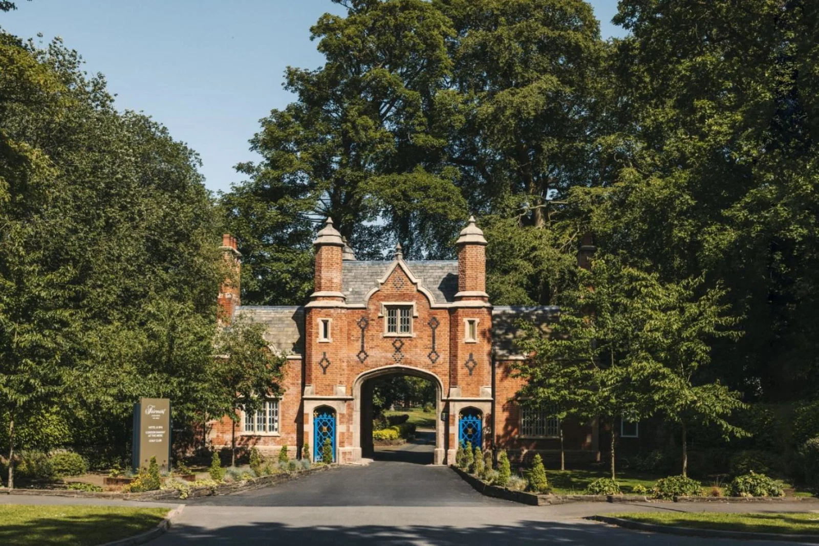 An ornate, 19th-century red-brick archway and gatehouse serves as the entrance to a luxury countryside estate, surrounded by mature green trees and manicured lawns under a clear blue sky. A modern hotel sign stands on the left edge of the driveway.