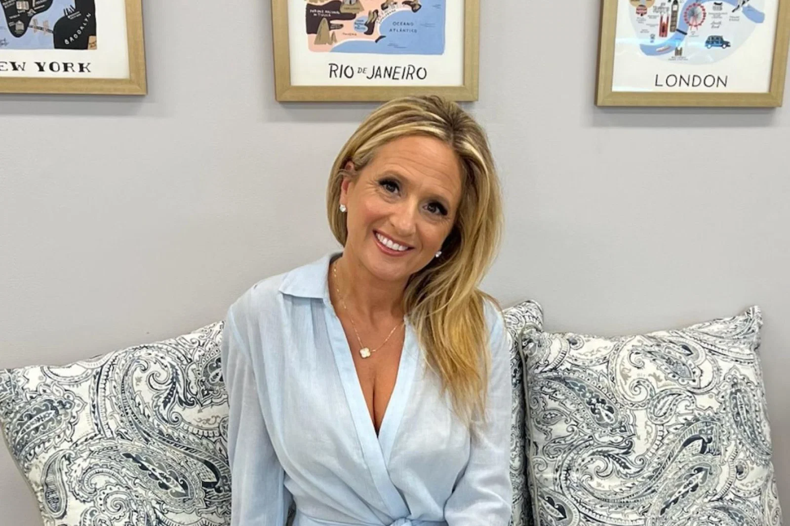 Woman with long blonde hair smiling, wearing light blue shirt, framed photographs in the background