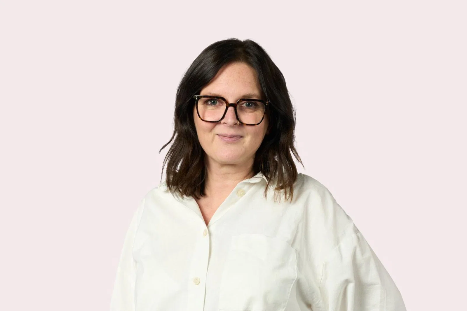 Headshot of brown haired woman with glasses and white shirt, light pink background,