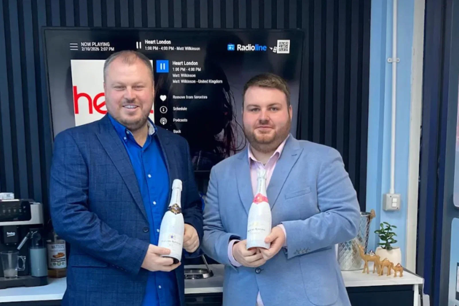 Two young white men standing in suits and holding champagne bottles