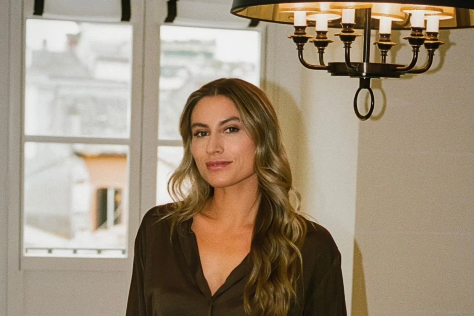White women with long, light brown hair smiling. There is a window allowing natural daylight and an overhead modern chandelier in the background. 