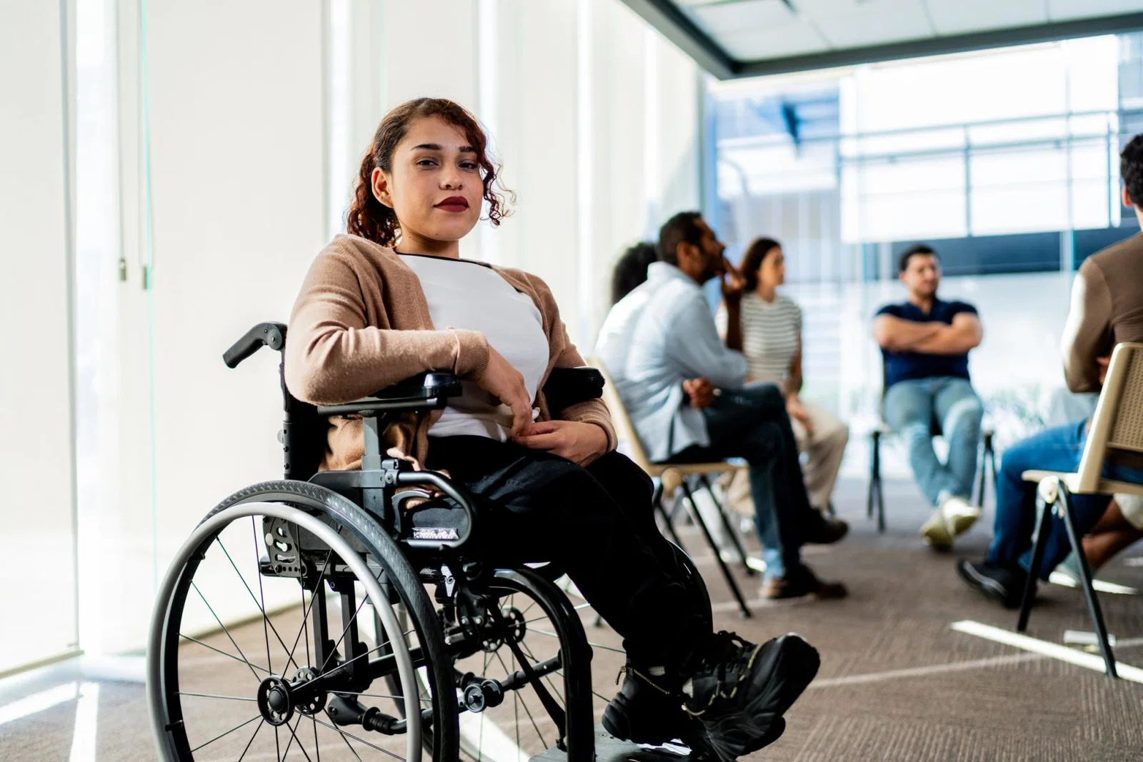Young woman using a wheelchair. She has curly brown hair, light brown skin and red lipstick. She is looking directly at the camera and there are a group of people speaking in a circle behind her.