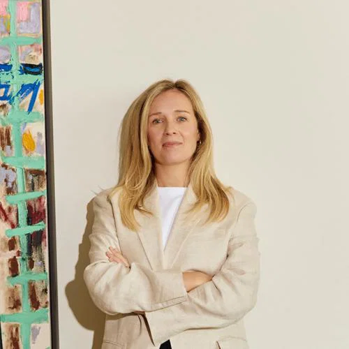 Blonde woman standing in front of white wall, to the left is a painting 
