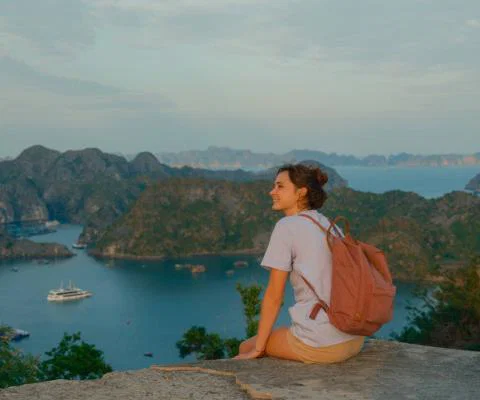 Brunette female solo traveller with backpack sitting on cliff edge overlooking scenic Vietnamese bay