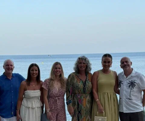 Six people, two men and four women, smiling in front of a beach 