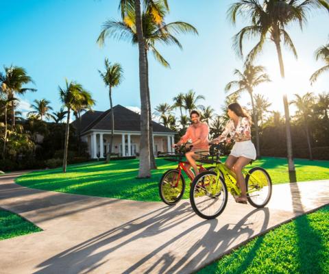 A couple cycling on paved path with grass and palm trees surrounding