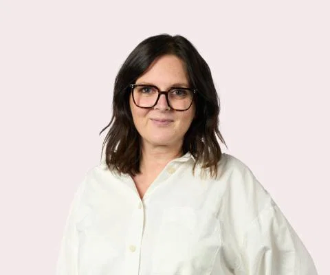 Headshot of brown haired woman with glasses and white shirt, light pink background,