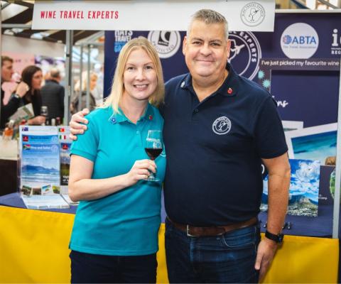 Blonde white woman and white man smiling at trade show, woman is holding glass of red wine 