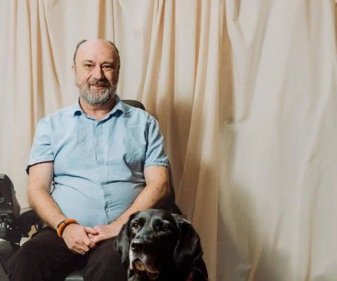 White man in his 50s, smiling in blue shirt, using wheelchair. A black dog is sitting to his right.  