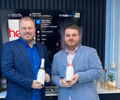 Two young white men standing in suits and holding champagne bottles