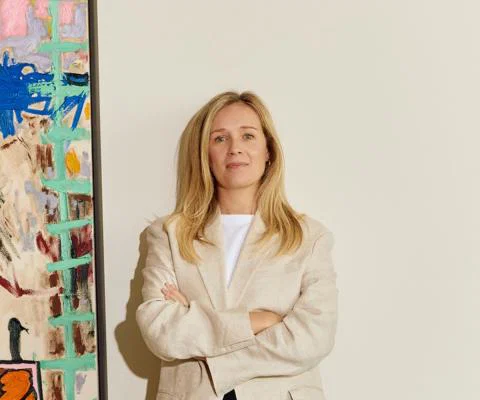 Blonde woman standing in front of white wall, to the left is a painting 