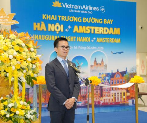 A man in a suit stands at a podium with microphones at a Vietnam Airlines event for the inaugural Hanoi to Amsterdam flight, with banners and yellow flowers in the background.