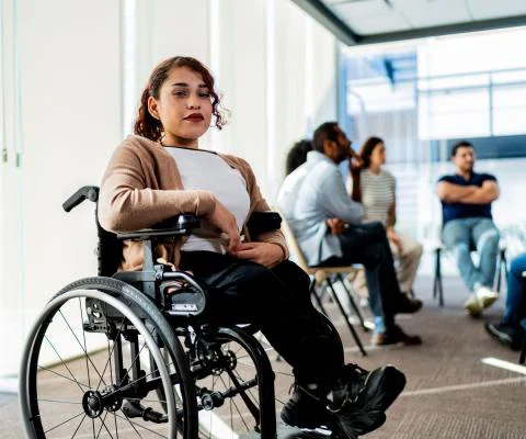 Young woman using a wheelchair. She has curly brown hair, light brown skin and red lipstick. She is looking directly at the camera and there are a group of people speaking in a circle behind her.