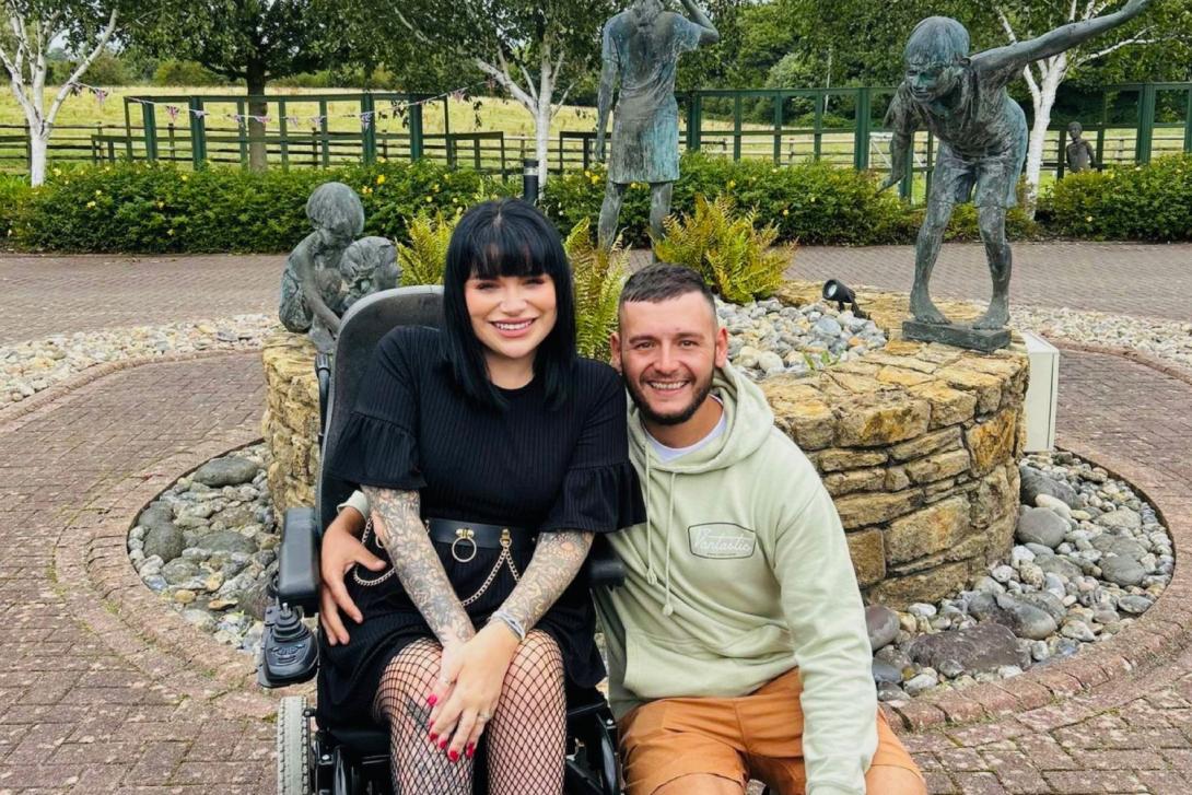 Young woman smiling with black hair, black t-shirt and skirt, using wheel chair. To her left is a smiling male partner, wearing green hoodie and orange shorts, kneeling down. They are in a park.