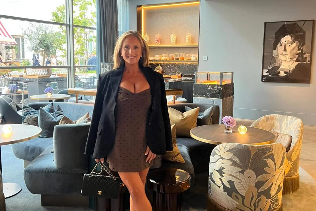 Woman with long blonde hair and brown dress with brown blazer smiling in brightly lit room