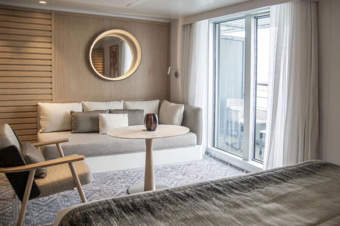 A luxury cruise ship suite wit a cream sofa, circular mirror on the wall, French windows leading onto a balcony