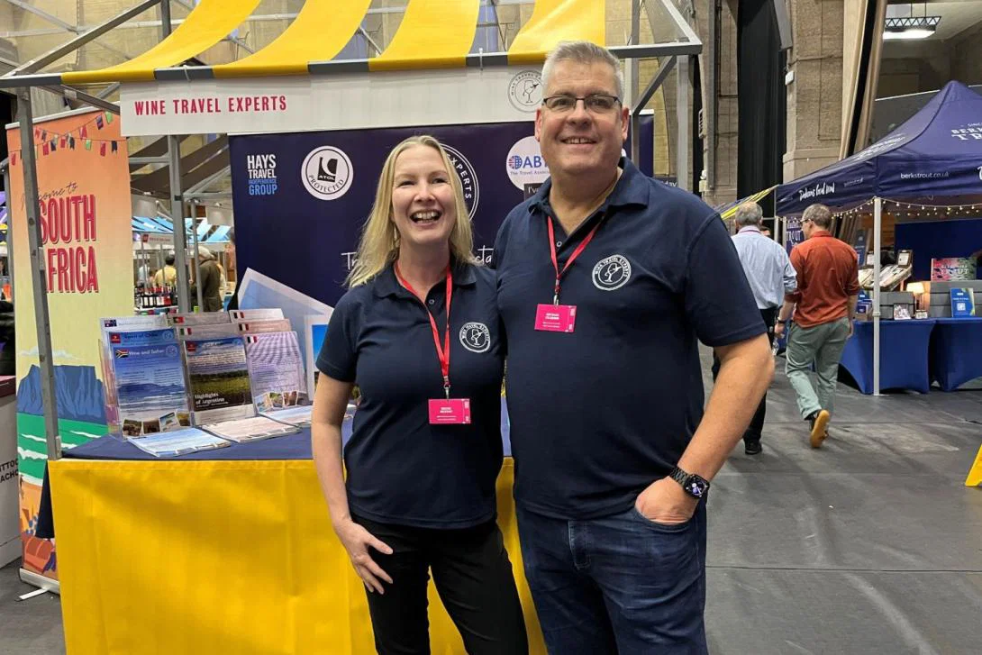 White blonde woman and man smiling in front of yellow wine stall, standing at trade show