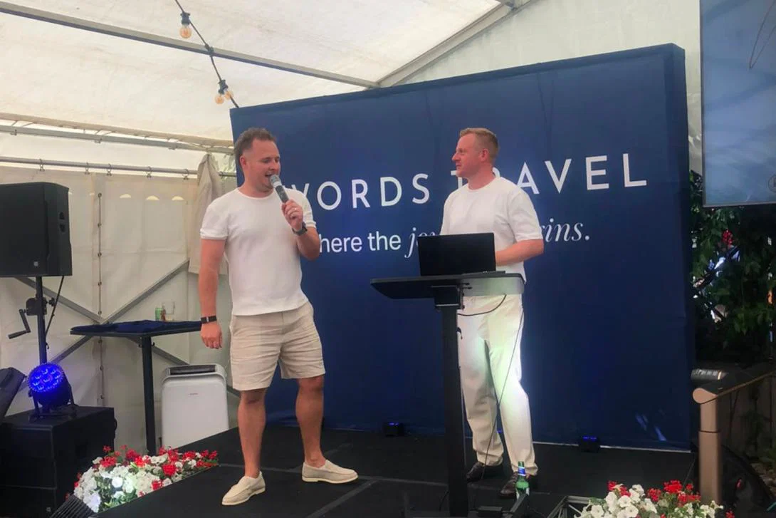 Two white men on small stage, wearing summery clothing and addressing an audience