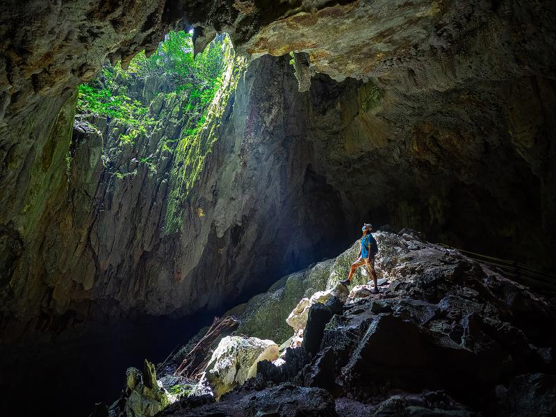 A man stands in cave with light shining on him through large opening above
