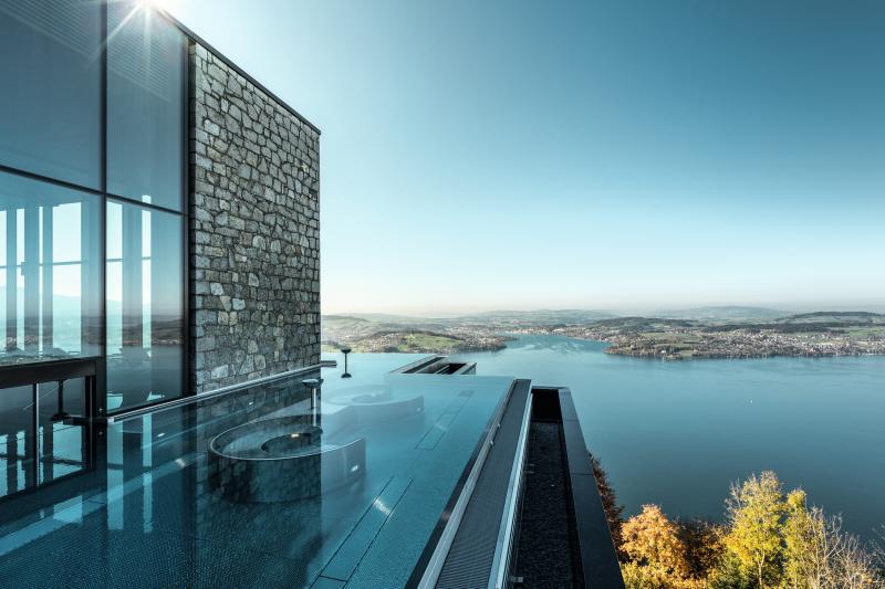A glass walled hotel overlooks a calm lake with trees surrounding 